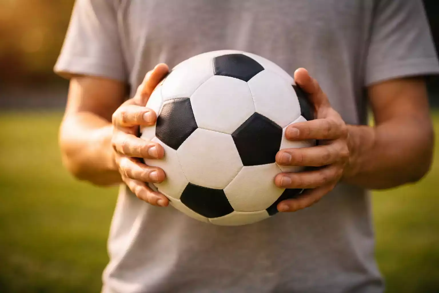 Manos abiertas sosteniendo un balón de fútbol con cuidado sobre un fondo verde suave